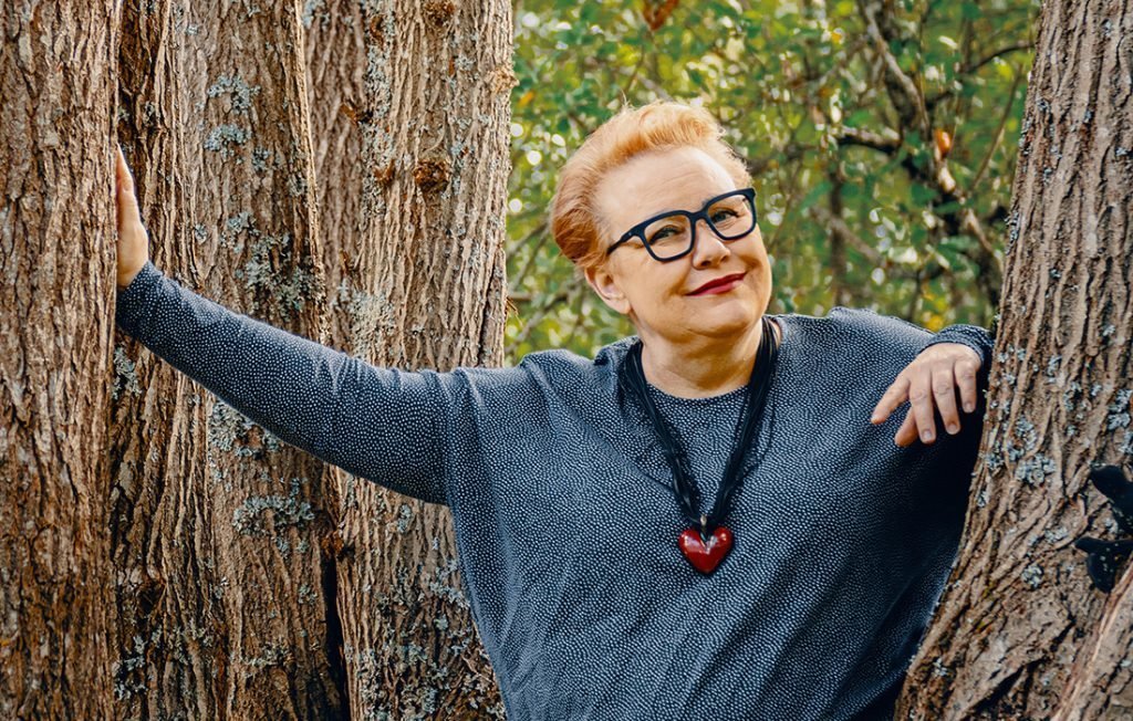 Sirpa Pietikäinen sinertävässä paidassa, sydänkoru kaulassa nojaa puuhun.